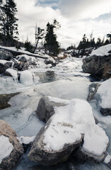 Vertical photo of river (stream) flowing in mountains with big snowy and frozen rocks.