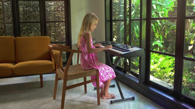 Blonde girl playing the piano in a beautiful modern home. Child is learning to play the electronic piano against the background of a glass wall and green garden.