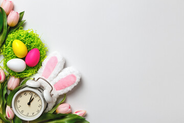Easter card. Multi-colored eggs in nests flowers tulips alarm clock with rabbit ears on a gray...