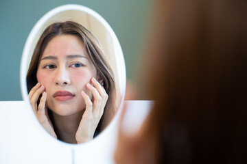 Asian young woman looking face skin in the mirror have a red rash on her face from cosmetic allergy