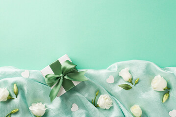 Top view photo of the nice white eustomas and green leaves with a small giftbox and confetti in shape of hearts scattered on the folds of the textured turquoise background copyspace