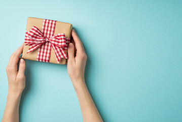 First person top view photo of valentine's day decorations young woman's hands demonstrating kraft paper giftbox with checkered ribbon bow on isolated pastel blue background with copyspace