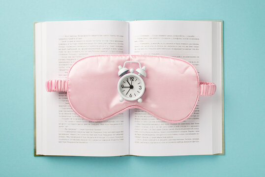 Top view photo of a white small clocks on a pink silk sleep mask situated in the middle of the open book on pastel blue isolated background