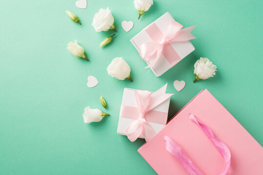 Top view photo of two small white gift boxes with cute silk pink bows white eustomas and small confetti in shape of hearts near the big pink shopping bag on the pastel turquoise background