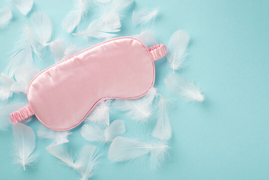 Top view photo of the big pink silk blindfold on the many white tender feathers scattered on the pastel sky blue isolated background copyspace