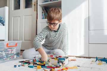Child is playing with construction toy blocks building a house in a white sunny play room.