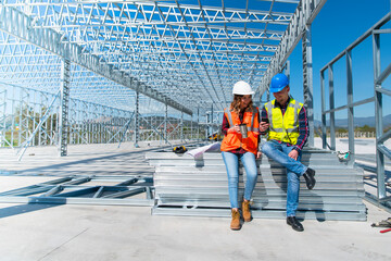 Cheerful construction workers smiling at the social media surfing while sitting. Beautiful young woman and handsome man, sitting in front of framework of the new building.