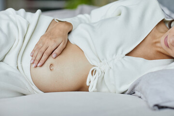 Close-up of young pregnant woman touching her belly lying on her bed in bedroom