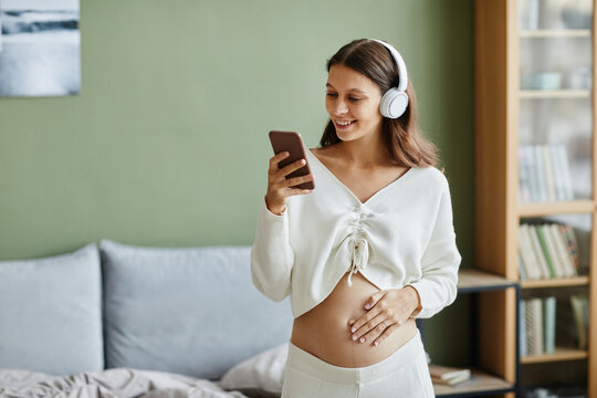 Young pregnant woman in wireless headphones enjoying the music on her mobile phone and dancing in the room