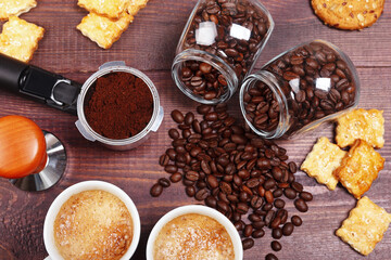 Two cups of espresso, crackers, cookies, holder with ground coffee, tamper and cans of coffee beans on wooden table.