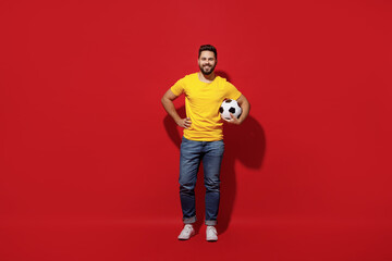 Full size body length fun young bearded man football fan in yellow t-shirt cheer up support favorite team hold soccer ball isolated on plain dark red background studio portrait. Sport leisure concept.
