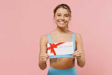 Young sporty athletic fitness trainer instructor woman wear blue tracksuit spend time in home gym hold gift certificate coupon voucher isolated on plain light pink background. Workout sport concept.