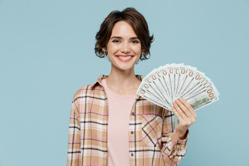 Young smiling happy woman 20s wearing casual brown shirt holding fan of cash money in dollar banknotes isolated on pastel plain light blue color background studio portrait. People lifestyle concept.