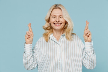 Elderly smiling blonde woman 50s wear striped shirt waiting for special moment, keeping fingers crossed, making wish, eyes closed isolated on plain pastel light blue color background studio portrait