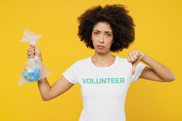 Young woman of African American ethnicity in white volunteer t-shirt hold in plastic bag Earth world globe show thumb down isolated on plain yellow background Voluntary greenhouse effect help concept
