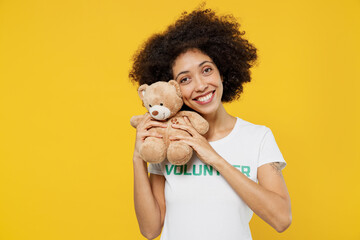 Young woman of African American ethnicity wears white volunteer t-shirt hold hug teddy bear plush toy isolated on plain yellow background. Voluntary free work assistance help charity grace concept