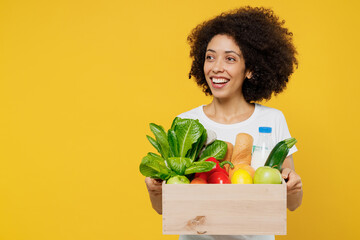Young happy smiling amazed woman of African American ethnicity wears white t-shirt hold box with vegetarian vegan food organic vegetables look aside on workspace isolated on plain yellow background - obrazy, fototapety, plakaty