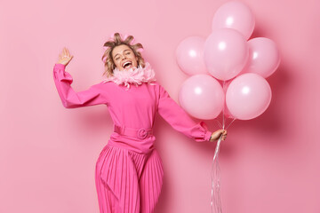 People fun and celebration concept. Carefree glad woman wears long dress applies hair rollers celebrates birthday holds bunch of helium balloons isolated over pink background dances with joy