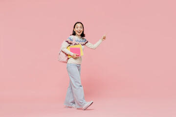 Full body happy teen student girl of Asian ethnicity in sweater hold backpack point index finger aside on workspace area isolated on pastel plain light pink background Education in university concept