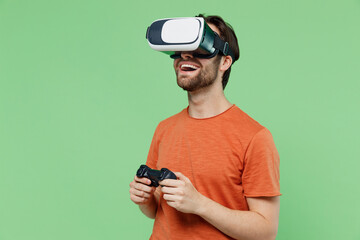 Young happy fun cool man 20s wear casual orange t-shirt hold in hand play pc game with joystick console watching in vr headset pc gadget isolated on plain pastel light green color background studio.