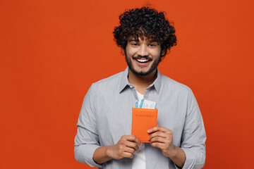 Fascinating smiling joyful fun charming happy young bearded Indian man 20s years old wears blue shirt hold passport boarding tickets looking camera isolated on plain orange background studio portrait.