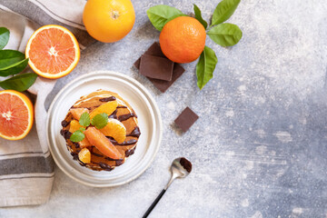 Celebrating Pancake day, healthy breakfast. Delicious homemade american bananas pancakes with chocolate and red orange on grey stone background. Flat lay, top view. Copy space.