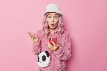 Shocked pink haired Asian woman stares bugged eyes with disbelief gets strange message on smartphone wears fur coat and panama stands against rosy studio background. Human reactions concept.
