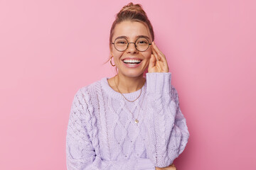 Portrait of pleasant looking woman smiles broadly shows perfect white teeth keeps hand on rim of spectacles dressed in knitted sweater isolated over pink background. People and positive emotions