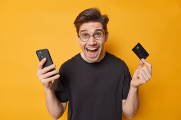 Positive handsome European man holds mobile phone and credit card makes online shopping or money...