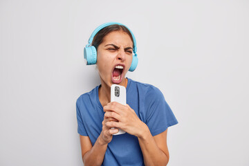 Carefree dark haired female singer keeps mouth widely opened listens music via headphones holds smartphone as if microphone enjoys loud sound dressed casually isolated over white background.