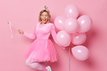 Obraz na płótnie Canvas Upbeat positive woman wears crown jumper and pleated skirt dances carefreeholds bunch of helium balloons and magic wand enjoy party celebratio isolated over pink background. Holiday concept.
