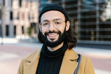 Portrait of positive young hipster man smiling at camera - Handsome trendy bearded guy with hat and glasses standing outdoors - Fashion and trendy business people concept