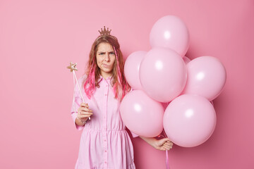 Obraz na płótnie Canvas Frustrated unhappy long haired young woman holds bunch of inflated balloons and magic wand being angry with someone wears dress and crown poses against pink background doesnt like something.