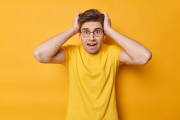 Shocked worried adult dark haired European man grabs head thinks about deadline looks stressed wears casual t shirt isolated over yellow background. People human reactions and emotions concept - obrazy, fototapety, plakaty