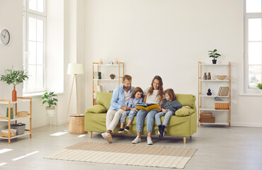 Happy family spending time at home. Mum, dad and little kids reading book while sitting together on green sofa in room with light walls, beige rug and simple brown shelves in their big new house