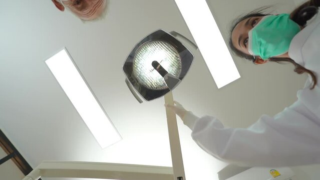 First person view of a young Asian female nurse and a senior Caucasian male dentist, in face masks, turned on the lights and doing a dental work for a patient laying on a chair with dental instruments
