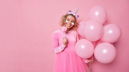 Obraz na płótnie Canvas Positive dreamy young woman wears festive clothes looks away has cheerful expression applies beauty patches wears unicorn headband holds bunch of inflated balloons isolated over pink background