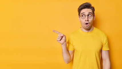 Impressed stunned adult man points finger away demonstrates awesome advertisement holds breath cannot believe his eyes wears round transparent glasses and casual t shirt isolated over yellow wall