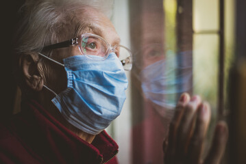 Old woman portrait at the window. Grandmother with medical mask staying home alone during...