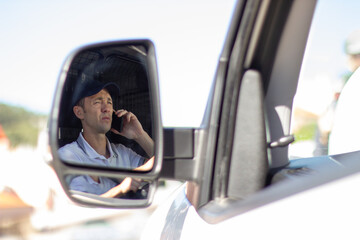 Deliveryman talking on mobile phone in car. Mid adult man in white T-shirt driving. Photo from mirror. Using delivery app. Work, delivery service, shipment concept