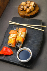 Rolls on a black carved board next to soy sauce and ginger and baked mushrooms.