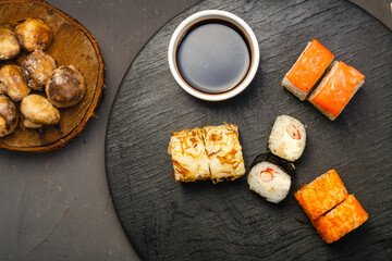 Rolls mini set on a round board next to sticks, soy sauce and baked champignons on a black background.