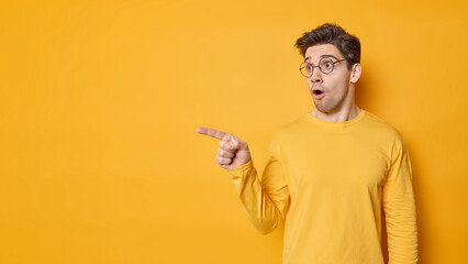 Stunned dark haired young man points away feels impressed shows stunning advertisement wears round spectaces and jumper isolated over vivid yellow background blank copy space for your promotion