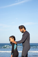 Naklejka na ściany i meble Happy bearded father and daughter near ocean. Mid adult man and little girl in surfing suit standing near water on cloudy day. Helping button up costume. Family, leisure, active lifestyle concept