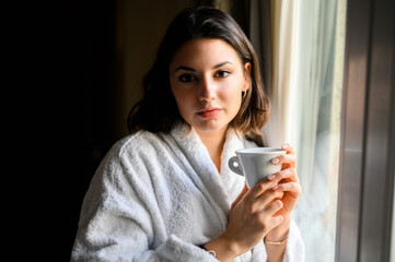 Young woman wearing a bathrobe and drinking a coffee near a window in a hotel room