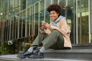 Outdoor shot of happy curly haired millennial girl wears fashionable clothes sits on stairs near modern shopping centre reads message on smartphone enjoys recreation time. Modern technologies - Powered by Adobe
