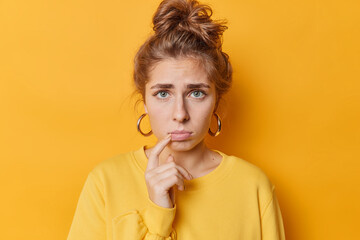 Portrait of good looking frustraed young woman looks sadly purses lips has combed hair wears earrings and casual jumper isolated over yellow background. Negative emotions and feelings concept