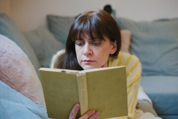 A woman lying on the couch reading a book - obrazy, fototapety, plakaty