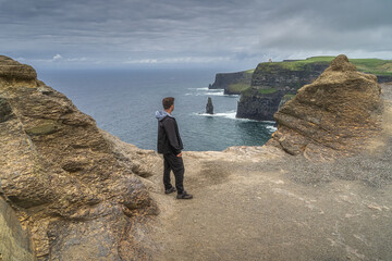 Man standing on the edge of iconic Cliffs of Moher and looking at sea stack underneath, popular...