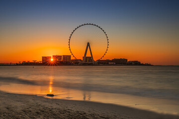Dubai beach at sunset. Jumeirah Beach Residence with the world largest Ferris wheel. Sunbeams between the houses. Ain Dubai at sunset with a blue orange sky